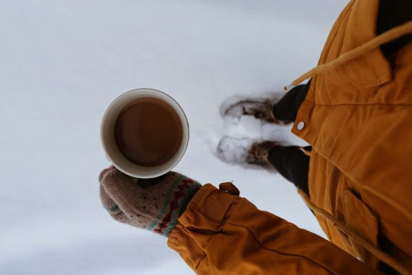 Person holding hot tea in hand with mittens and winter clothing on, in the snow. Photo by Sabina Sturzu on Unsplash.