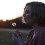 Woman blowing out the mouth at a dandelion flower. Photo by Havilah Galaxy on Unsplash