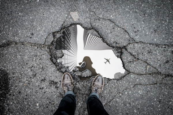 Reflection in puddle of person in city with airplane overhead. Photo by Marc-Olivier Jodoin on Unsplash