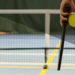 Person holding pickleball paddle and ball on court, half shaded. Photo by Aleksander Saks on Unsplash.